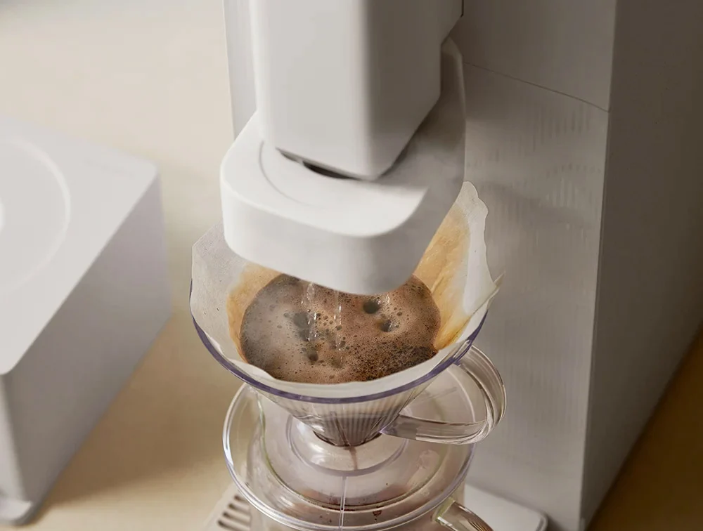 A close-up of STEAM 100 Barista water purifier dripping freshly brewed coffee into a glass carafe, with a white paper filter cone in the cone holder.